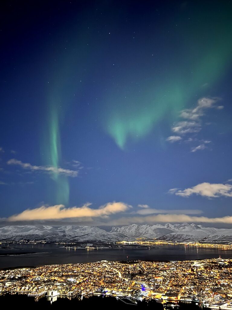 Northern lights swirling green over the fjord and city lights of Tromsø, Norway